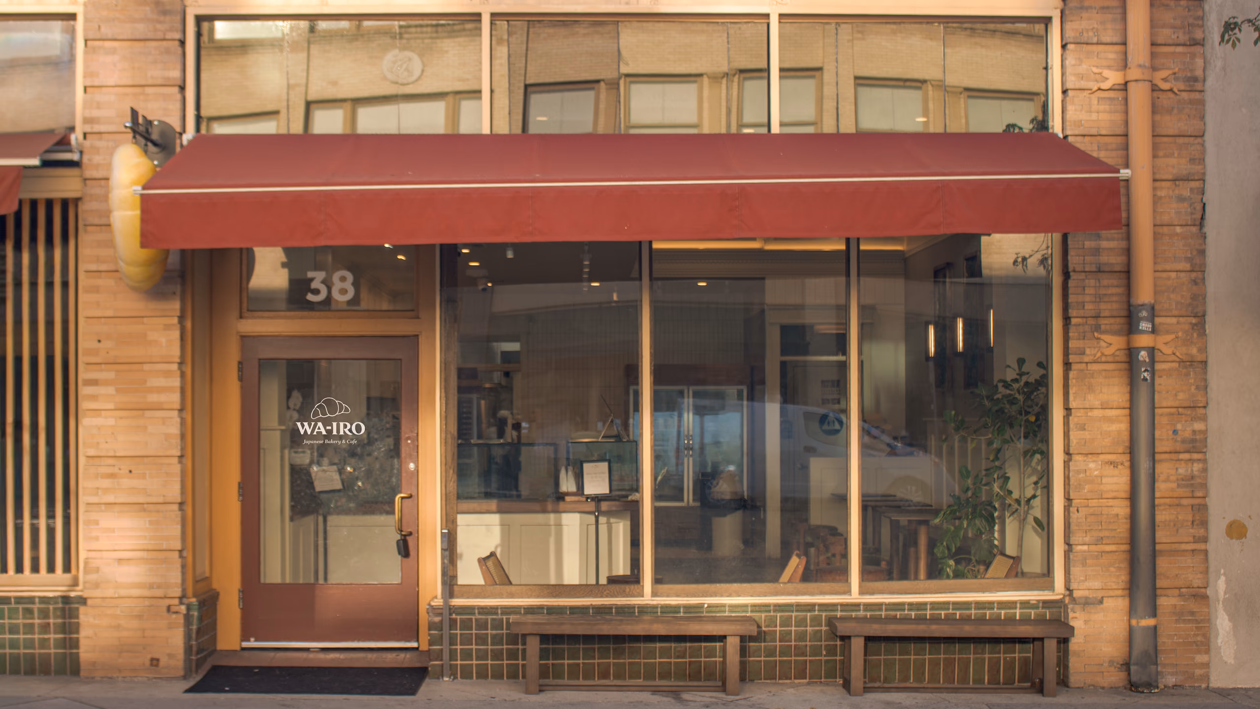 The Wa-Iro storefront seen square-on from the sidewalk: a deep red awning over tall windows, the street number 38 beside a glass door carrying the WA-IRO croissant logo, wooden benches below the windows, and the sculptural hanging croissant at the left edge.