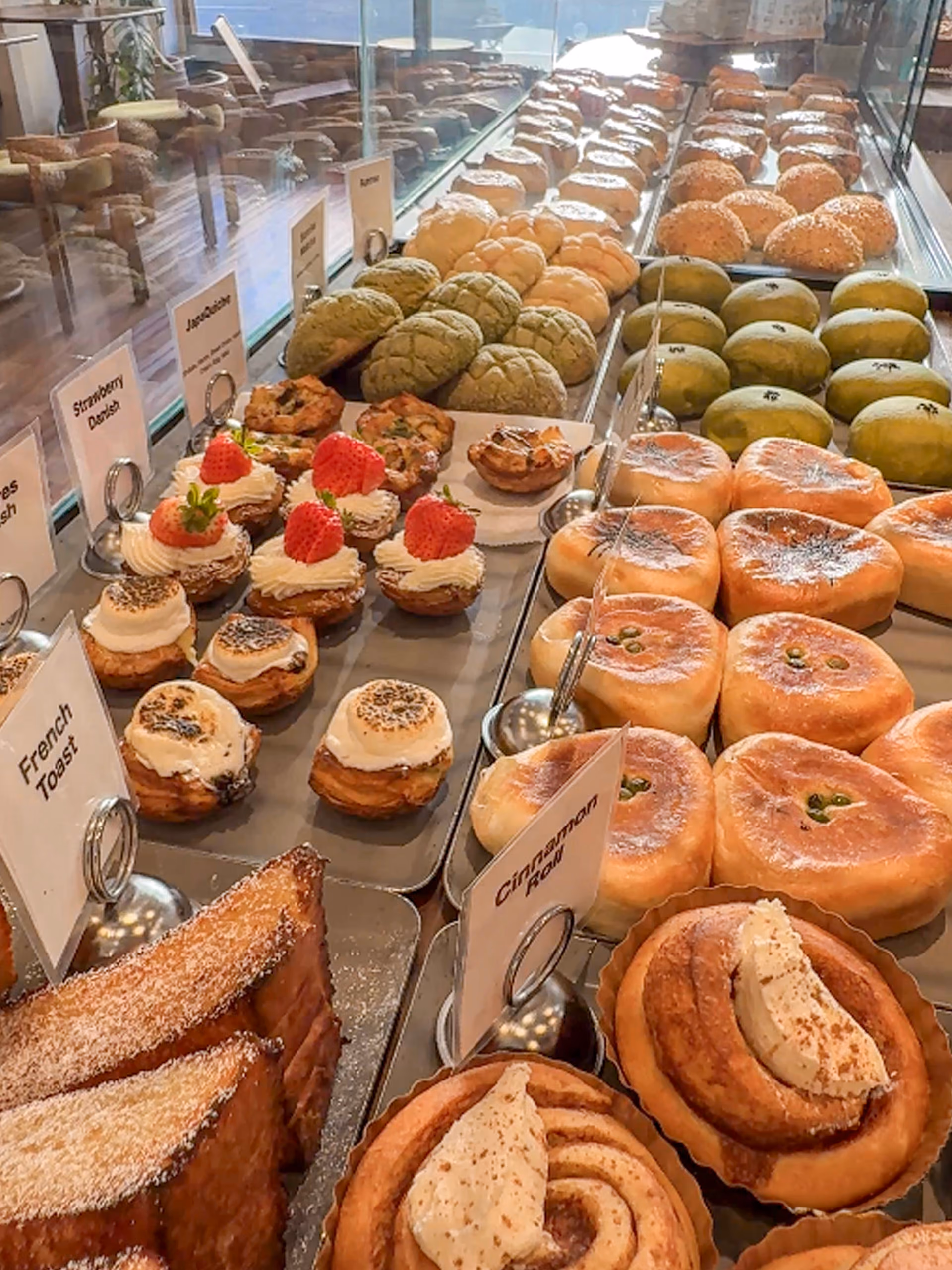 A bakery case seen down its length — thick-cut French toast, cinnamon rolls, cream buns, matcha melon pan, strawberry-topped danishes and marshmallow-topped pastries filling tray after tray.