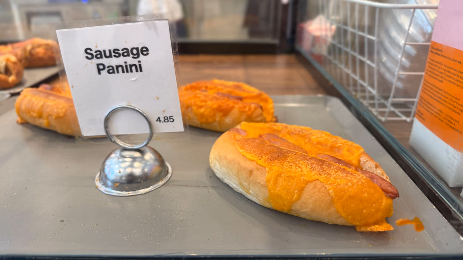 A Sausage Panini sandwich with melted cheese in the bakery case, a "Sausage Panini $4.85" price tag beside it.